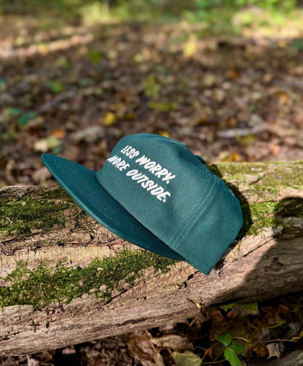 A dark teal baseball cap with white embroidered text reading 'ESS NO RRRRY WIR OUTSIDE.' rests on a mossy log in a forest setting.
